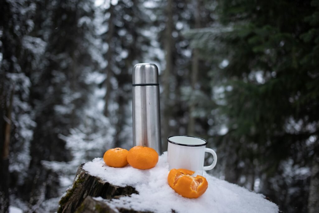 A serene winter scene featuring a thermos and fresh oranges on a snowy stump in a forest.